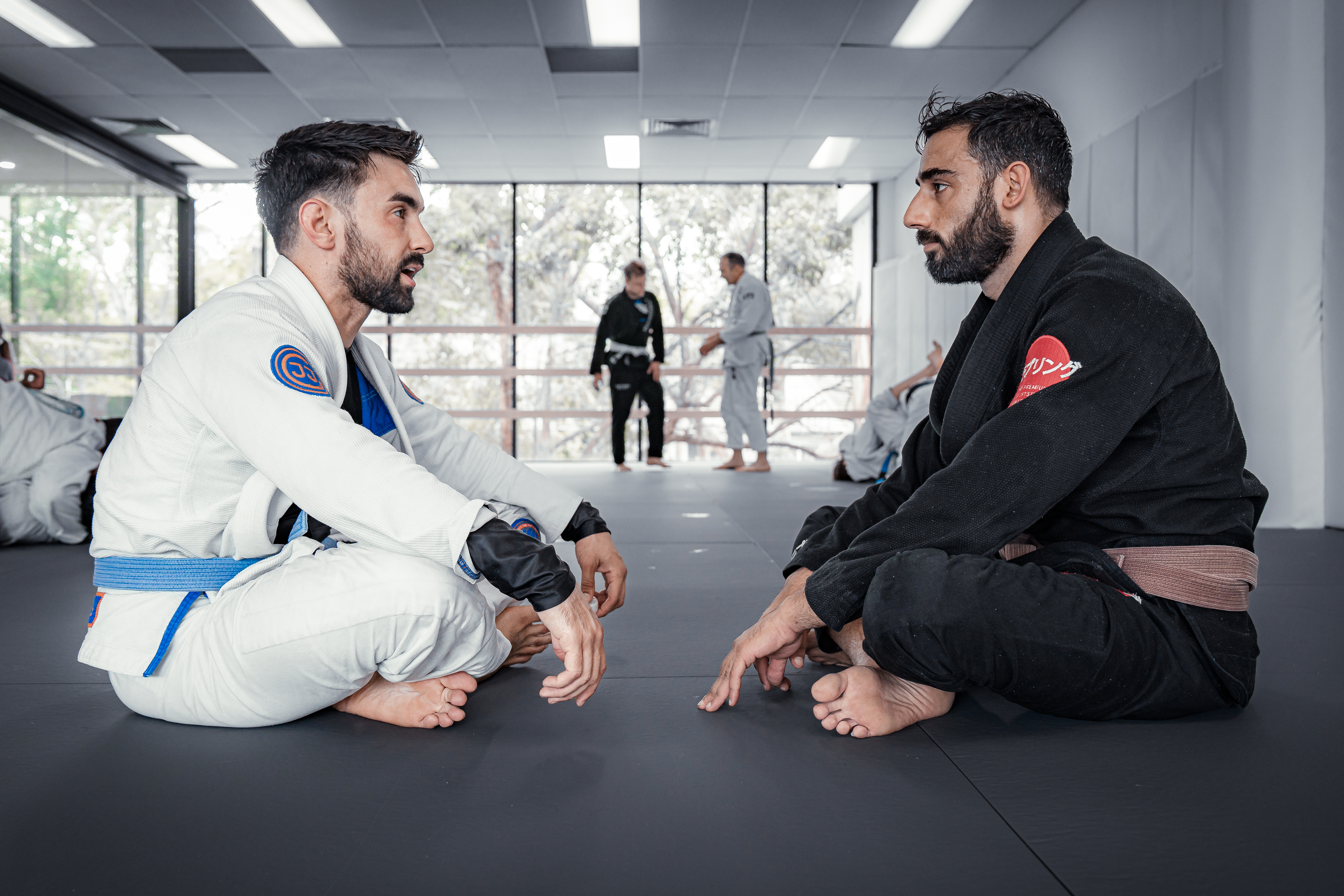 A junior and senior student seated in front of each other, discussing Brazilian Jiu Jitsu, in traditional Jiujitsu uniform known as the Gi